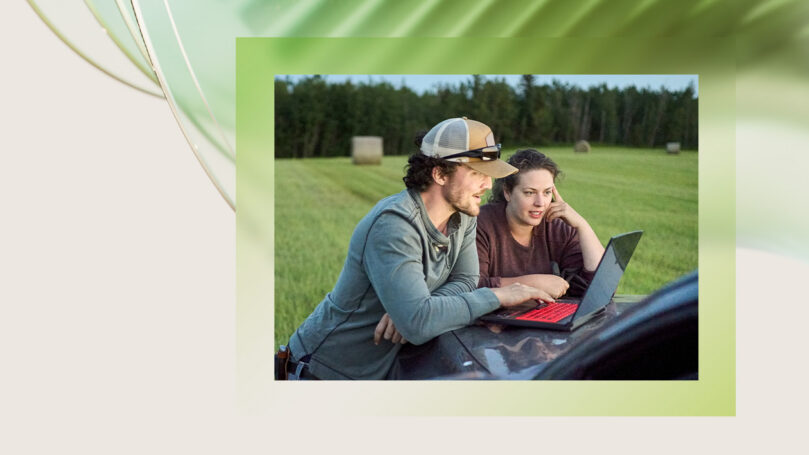 Two people looking at a laptop on the hood of a car parked in a hayfield.
