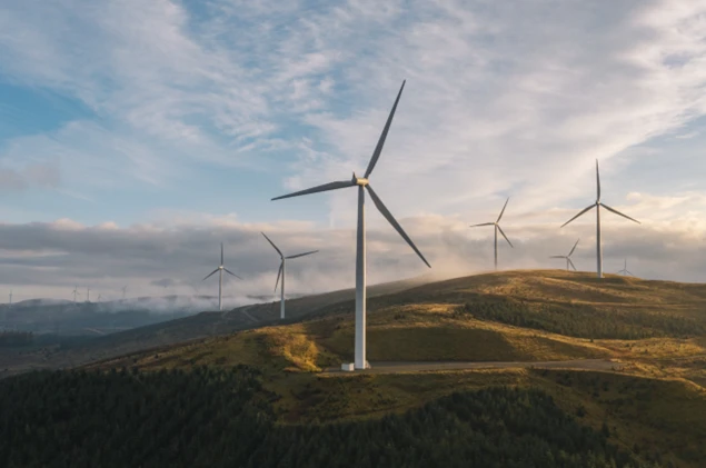 Wind turbines on rolling hills at sunrise, generating renewable energy above a forested landscape.
