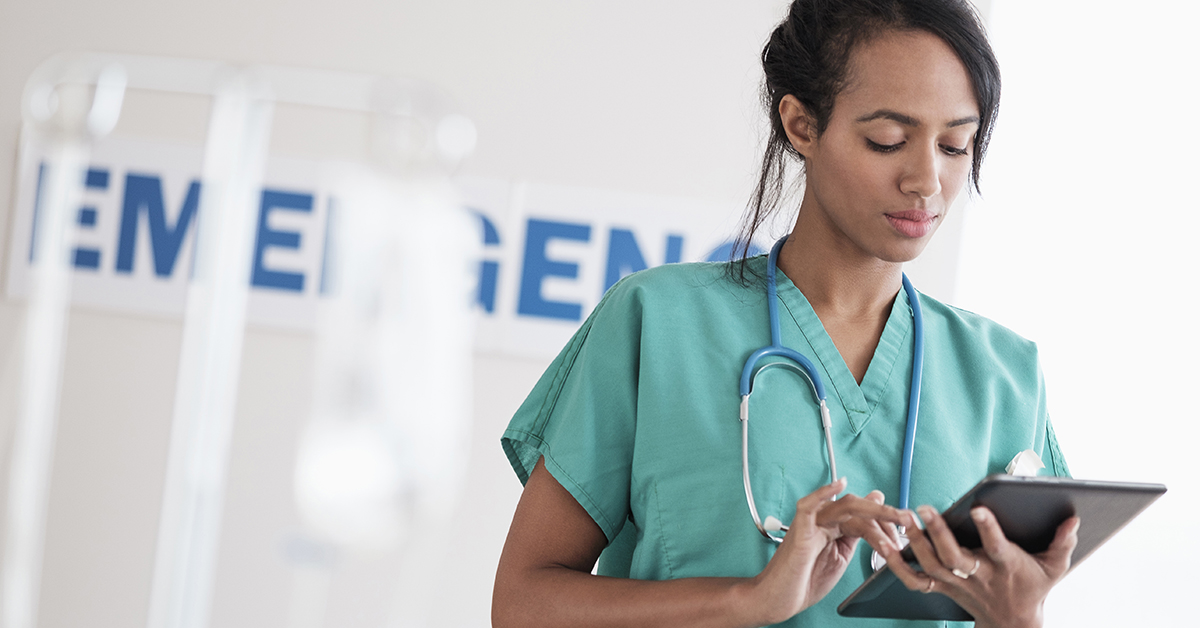 Healthcare professional in teal scrubs using a tablet, with a stethoscope around their neck, standing in a clinical setting with medical equipment in the background