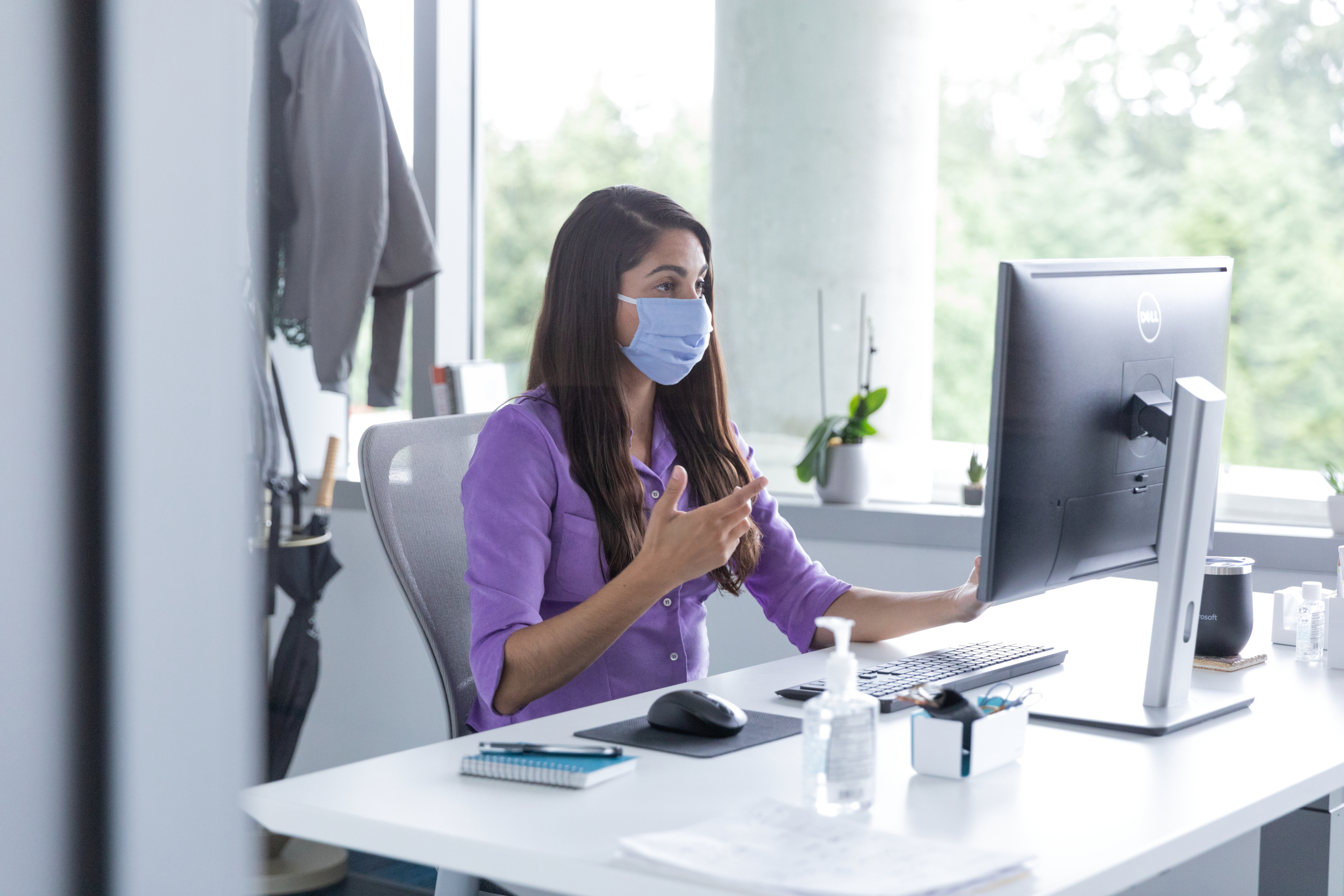 A female employee talking on a video call with hand sanitizer on her desk, socially distancing from others. Hybrid Workplace collection. Keywords: Hybrid Workplace, Hybrid Worksite, Return to Workplace, Return to Work, Return to Worksite, Return to Office, Return to Campus, Reopening, Back to work, COVID-19, COVID-19 policies, COVID, Coronavirus, Pandemic, Health, Health and Safety, Face Covering, Face Mask, Masks, Masks at Work, Masks in Workplace, Open Workspace, Open Workspace Policies, Social Distancing, Social Distancing Policies, Social Distance, Physical Distancing, Hand Sanitizer, Clean Office