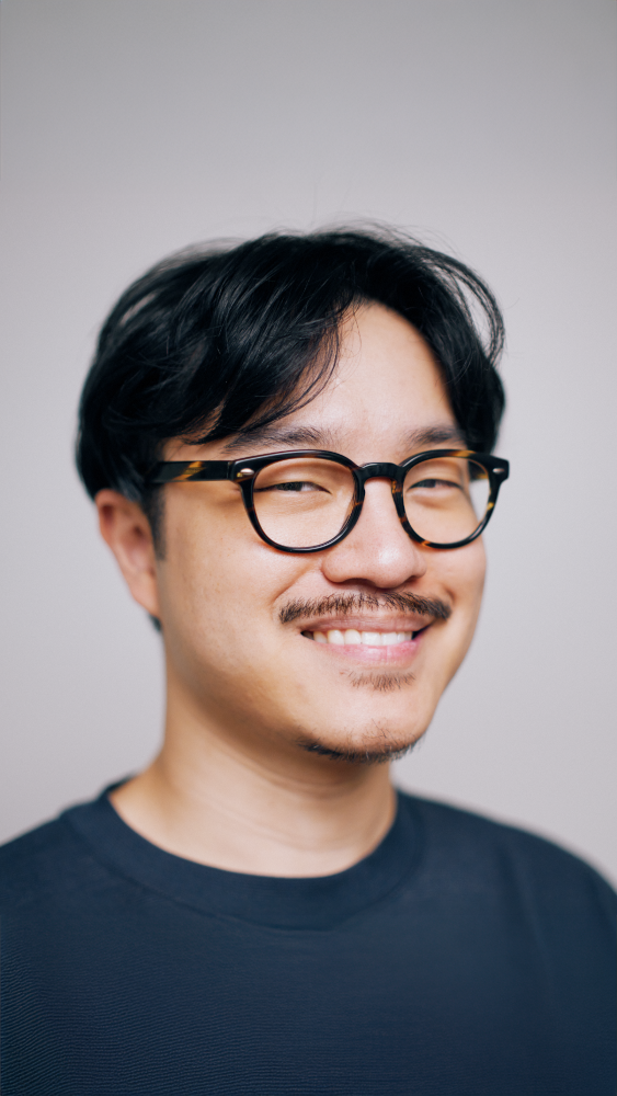 Person with short dark hair wearing glasses and a dark crew-neck shirt against a plain light gray background