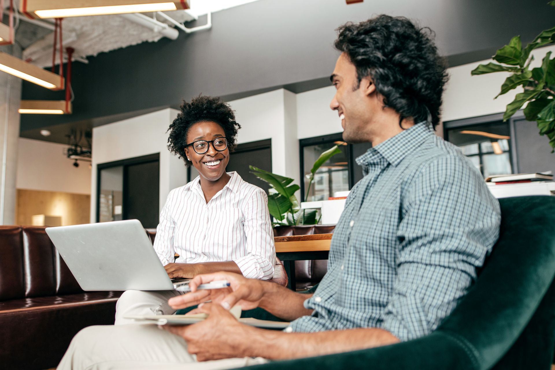 two people chatting looking at each other. one is holding a laptop and another is holding a notepad with a pen