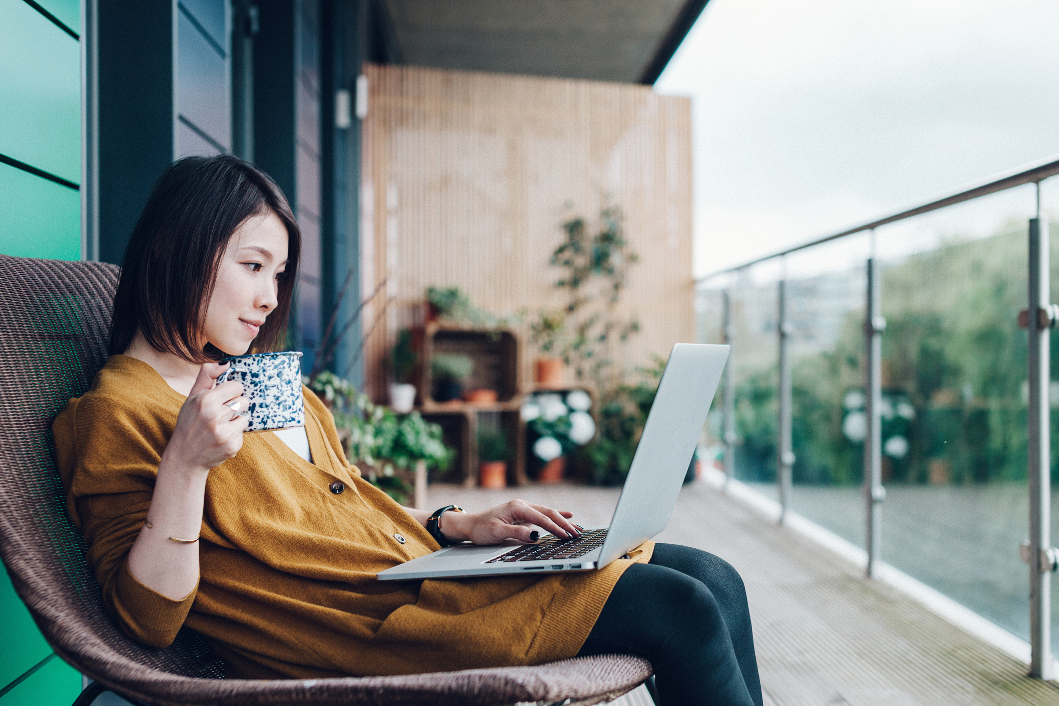 A female freelance remote worker sits on an apartment balcony with a latop and cup of coffee.