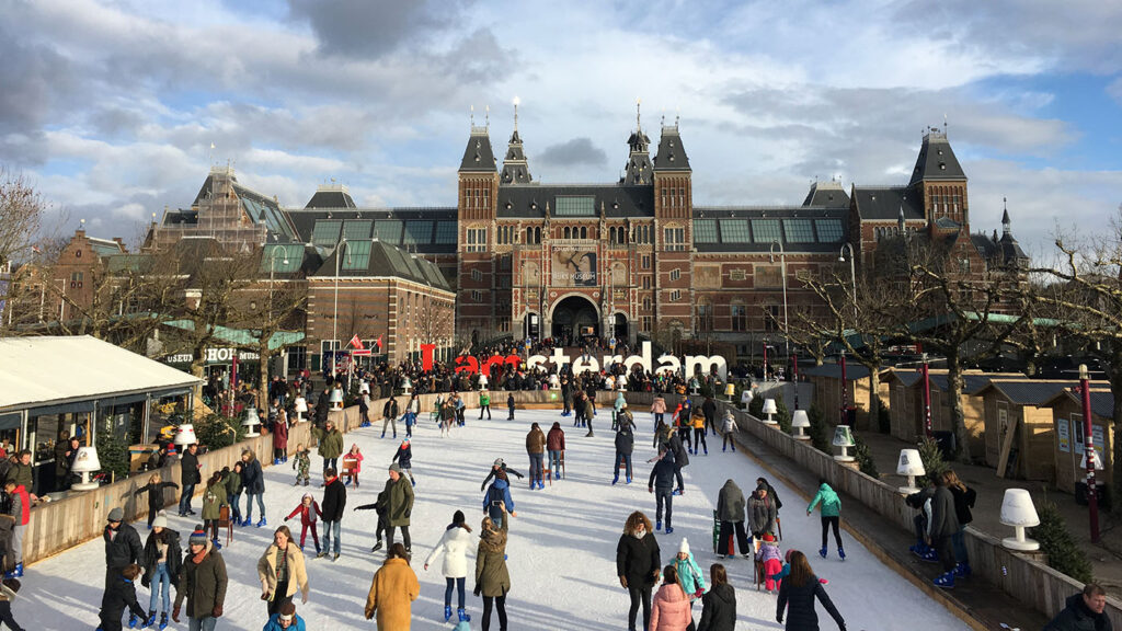 Photo of the Rijksmuseum, a huge grand building with a combination of gothic and renaissance architecture. In front of the museum are large letters spelling out ‘I Amsterdam’, surrounded by people ice skating.