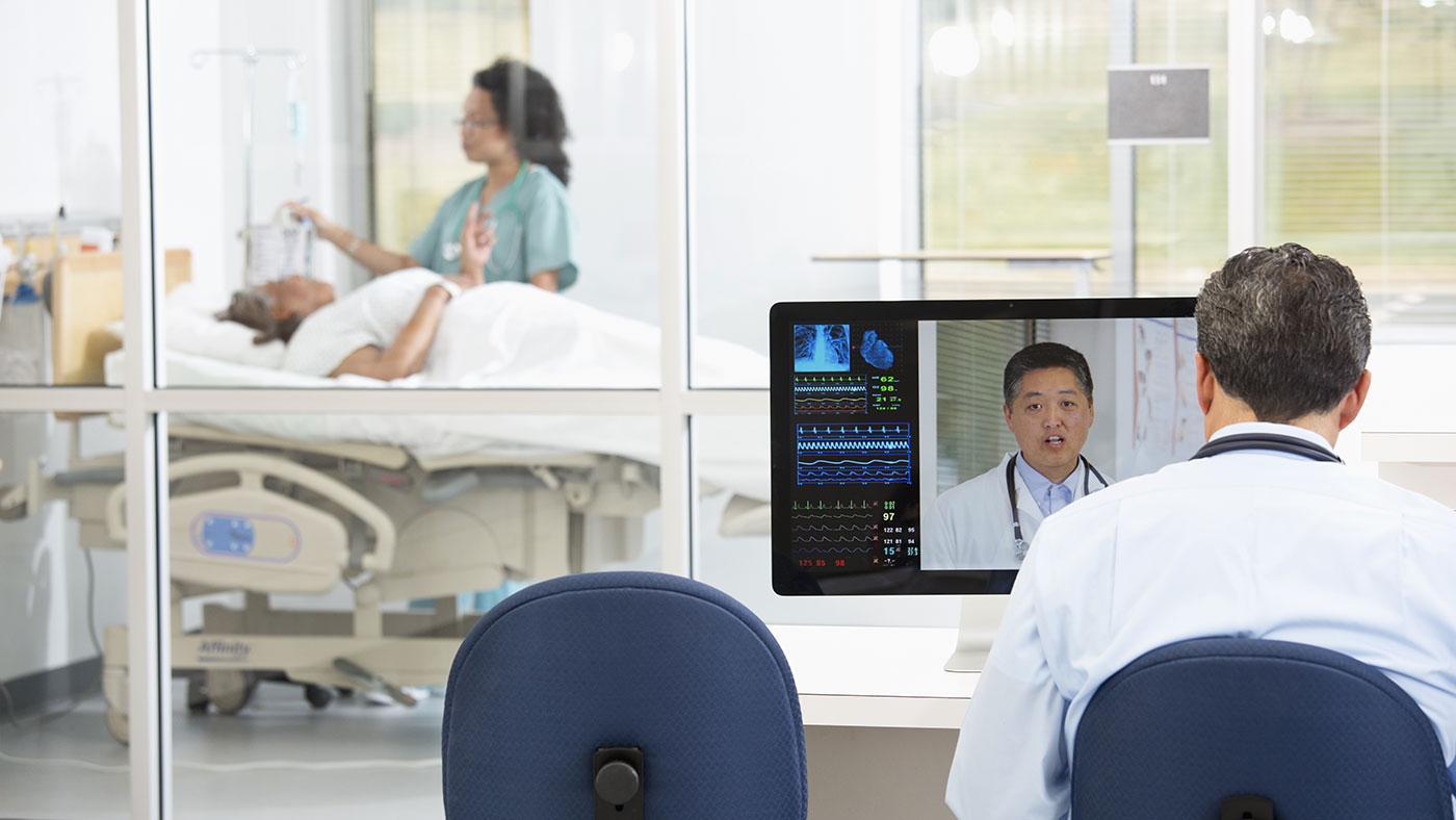 patient in a hospital bed being attended to by nurse with a doctor outside the room on a video appointment