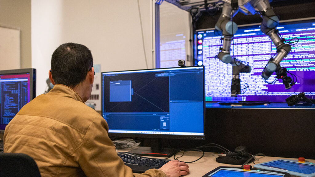 Researcher working on a computer screen with two robotic arms suspended from a frame with a large screen of code behind