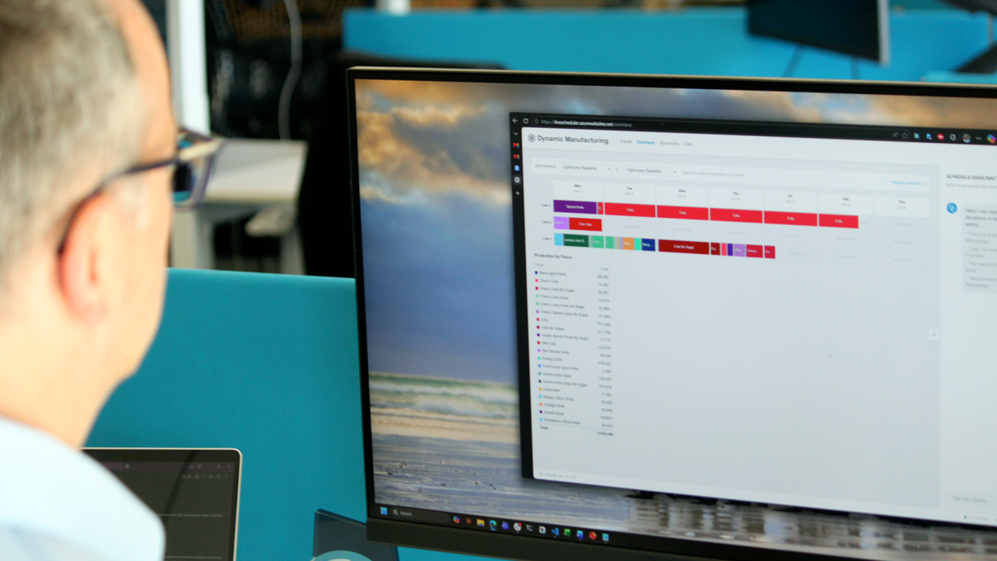 Person viewing a desktop monitor displaying a “Dynamic Manufacturing” dashboard with a color-coded production schedule timeline and resource breakdown, showing optimized scheduling results across multiple production lines.