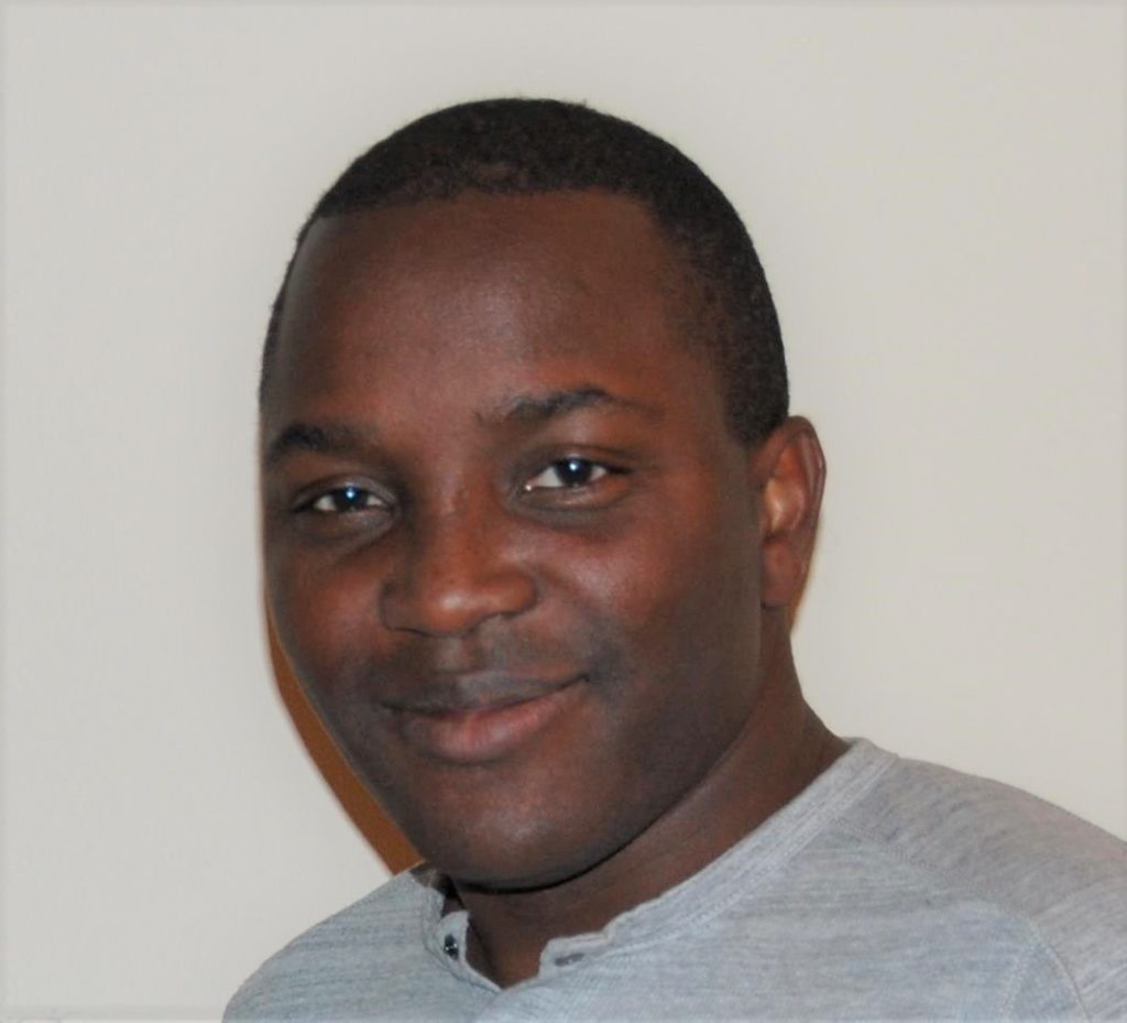 A profile headshot of Joe Dadzie, wearing a grey shirt against a cream-colored wall.
