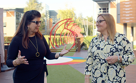 Two women talk outside a corporate office building with art in the background.