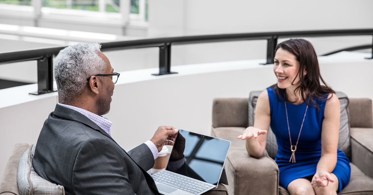 Image of two office workers talking. One has a laptop open.