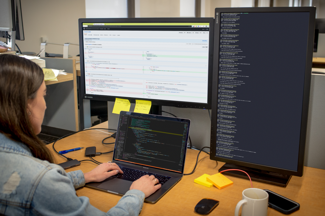 Developer coding at her workspace in an enterprise office using Visual Studio for Mac on a multi-monitor setup.