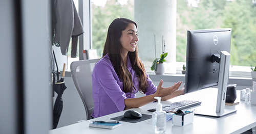 An image of a female employee talking on a video call with hand sanitizer on her desk, socially distancing from others.