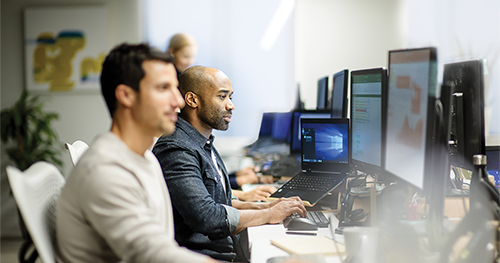 An image of two male coworkers sitting next to each other at along row of office desks, using their own desktop computers.