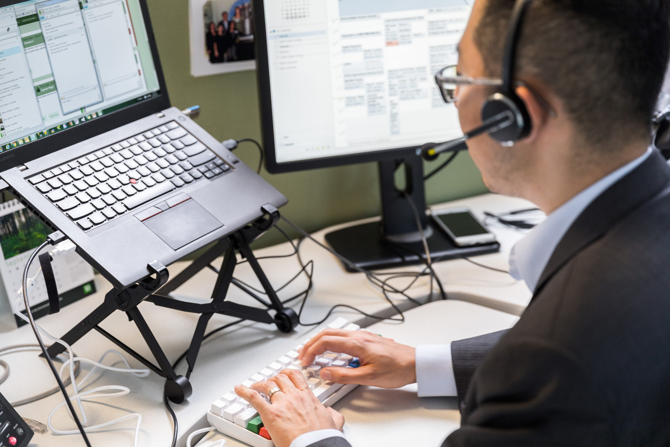 Male worker sitting at cubicle desk in financial office, wearing glasses and phone headset.