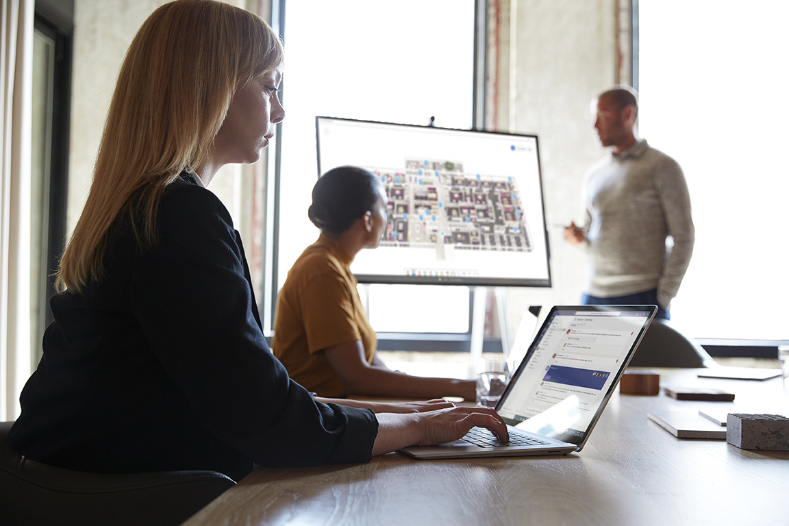 Three adults are collaborating in a conference room. One male is standing next to a Microsoft Surface Hub 50&rdquo; with Microsoft Whiteboard screen shown.