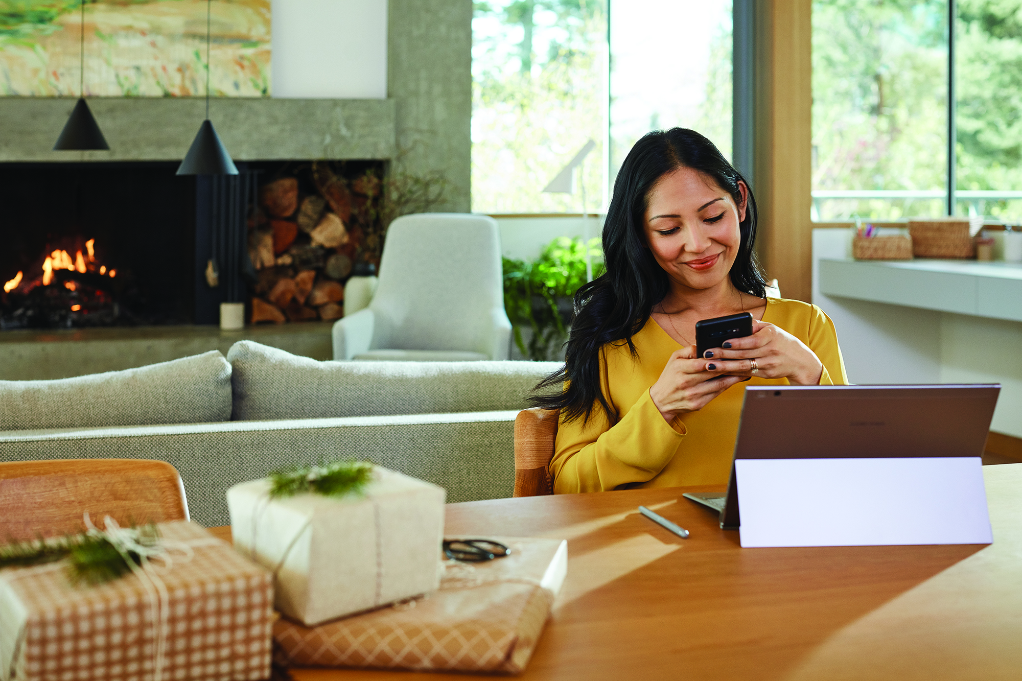 Image of a woman sitting at a table in front of a Surface, looking down at her phone. There is a couch and fireplace visible behind her.