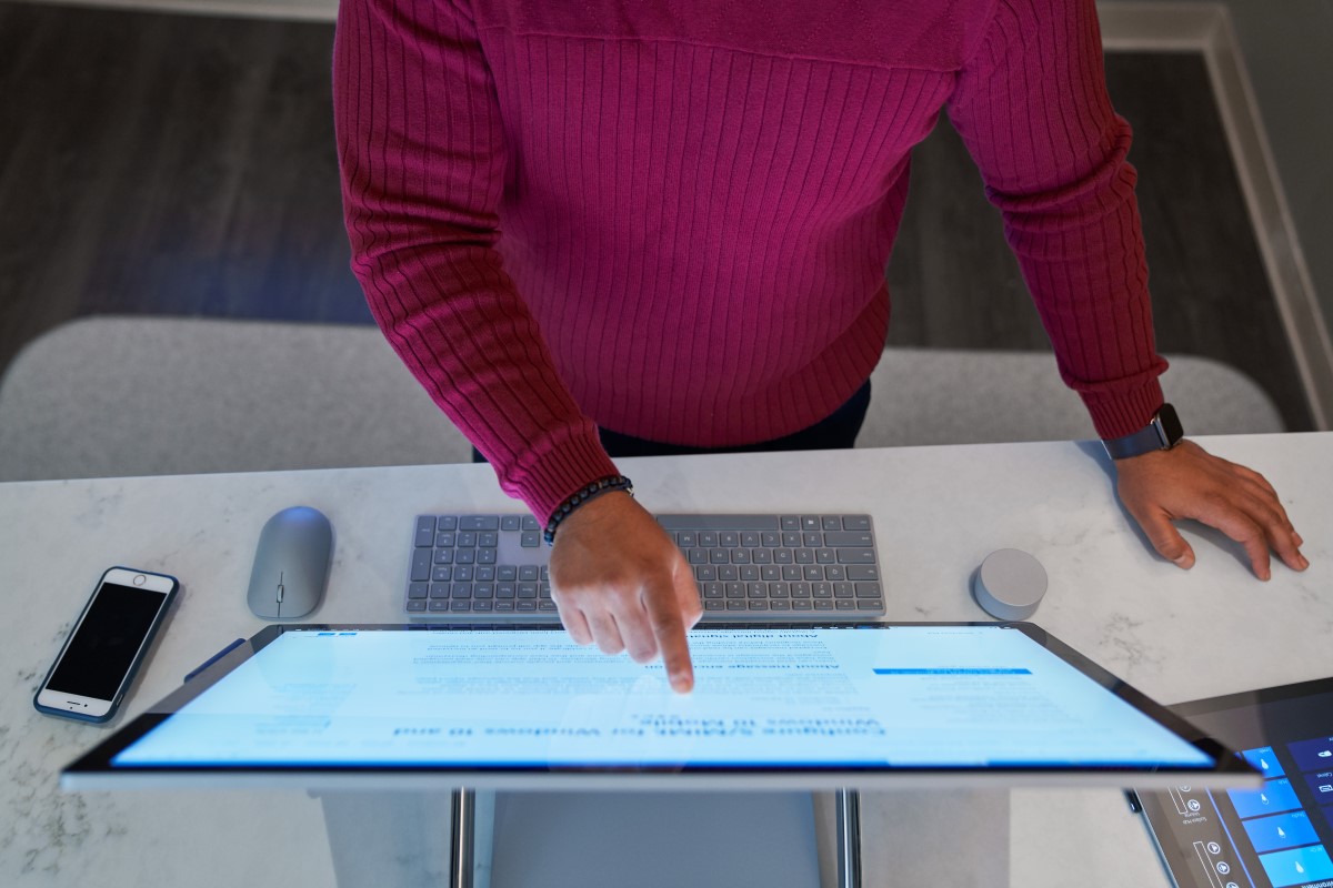 Top down view of a man wearing a dark red shirt working on a Microsoft Surface Studio with a phone next to him on the desktop along with a mouse, keyboard, and Surface Dial.