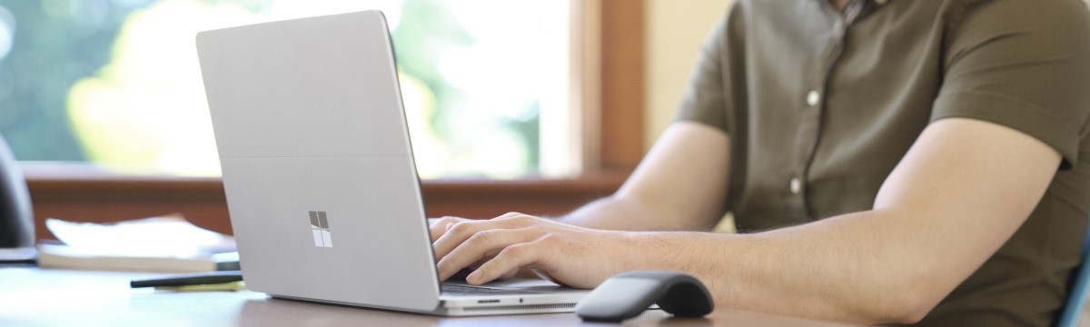 Male teacher sitting at a desk typing on a Surface Laptop Studio in laptop mode. A Microsoft Arc Mouse sits on the desk.