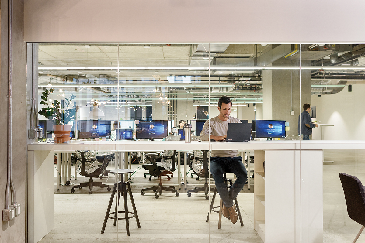 Male office worker seated at high desk in modern office, using Toshiba laptop (screen not visible). Rows of office workstations are in the background, each with desktop computers (some screens partially visible open to Windows 10 log in page).