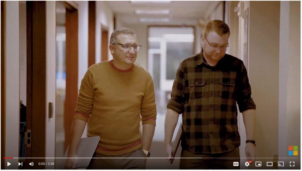 Two coworkers walk down office hallway carrying individual laptops. 