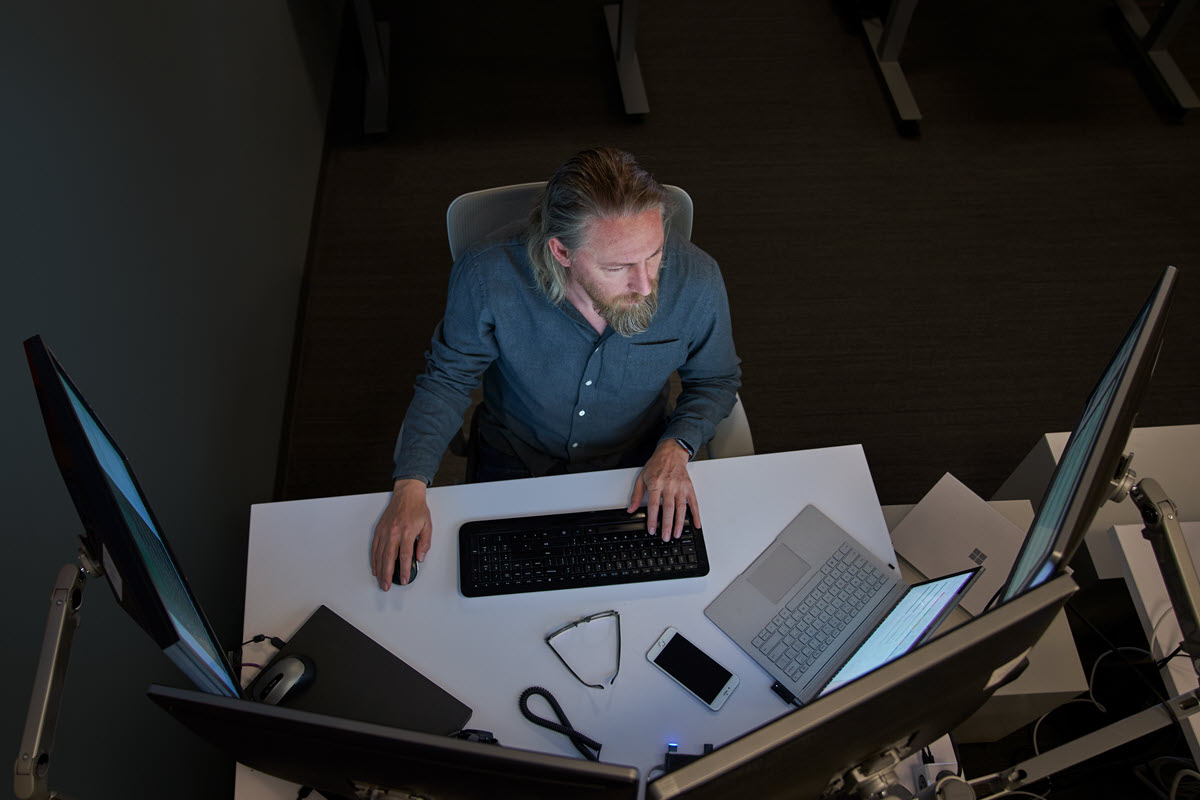 Top-down view of a bearded man in a gray/blue shirt seated at a desk working on a Surface laptop connected to three large monitors.