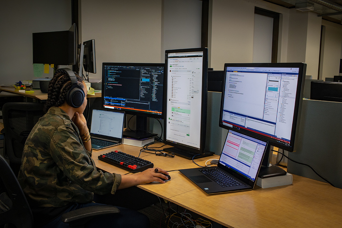 female developer wearing headphones; coding at her PC workspace in an enterprise office, using Visual Studio on a multi-monitor set up.