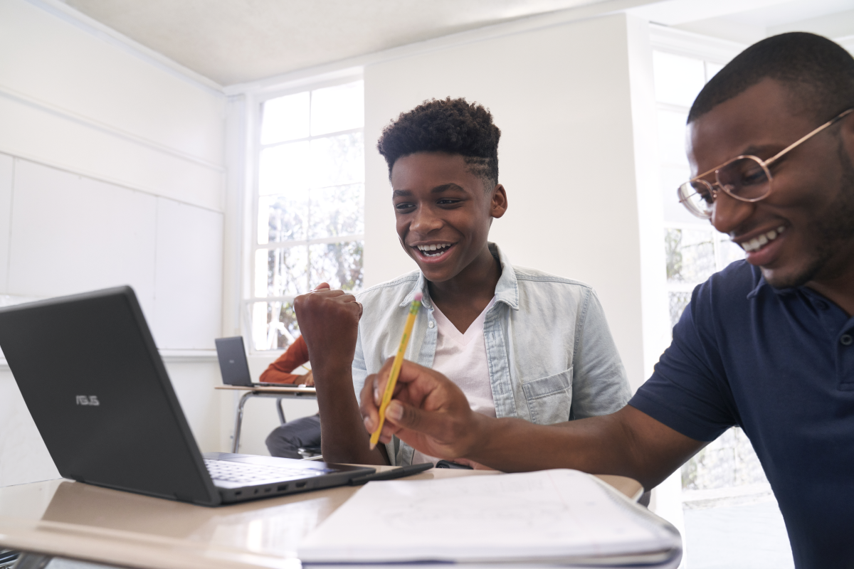 Excited male K-12 student sitting at a classroom desk using an ASUS BR1100 CKA device. Smiling male educator kneeling beside him.