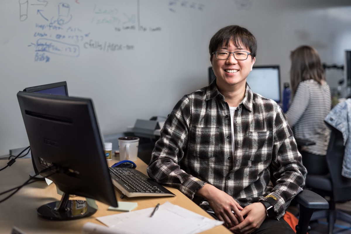 Portrait of male developer in glasses and plaid flannel shirt, smiling and facing camera. He is sitting at a desk in an office setting, with both hands on his lap. Keyboard and desktop computer visible, though screens are not shown. A female worker is shown seated at desk using computer in background (screen blurred). Whiteboard wall also shown in background with handwritten diagrams and formulas.