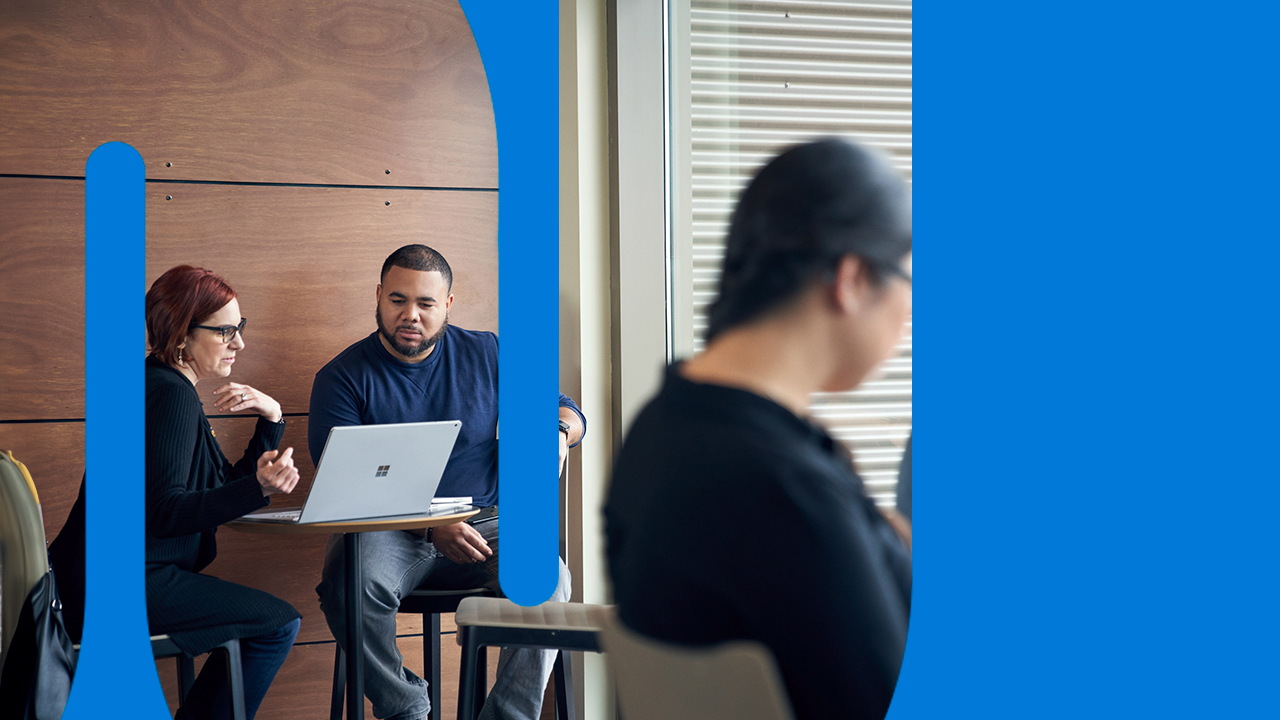 Man and woman meeting together and reviewing content on a Surface laptop at a high table with two blurred women in the foreground.