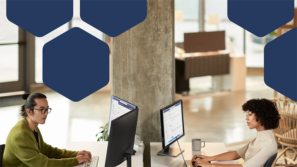 Photo of two employees collaborating during a Microsoft Teams meeting while working in an open office setting on dual monitors.