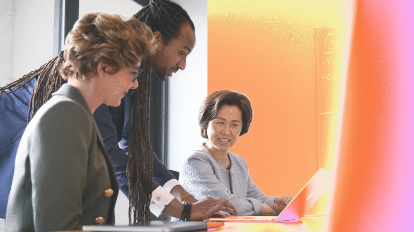 Three people collaborating at a desk, with two women sitting and one man standing in between them