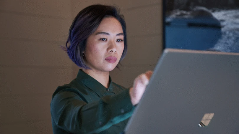 Side profile of a woman wearing a dark shirt in a dim office reaching up and working on a Microsoft Surface Studio.