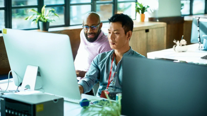 Two men look at a computer screen in a sunny office.