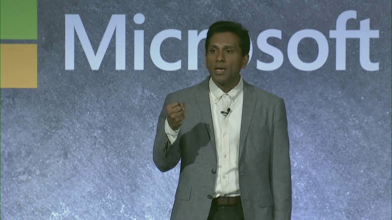 Joseph Sirosh wearing a suit and tie standing in front of a sign