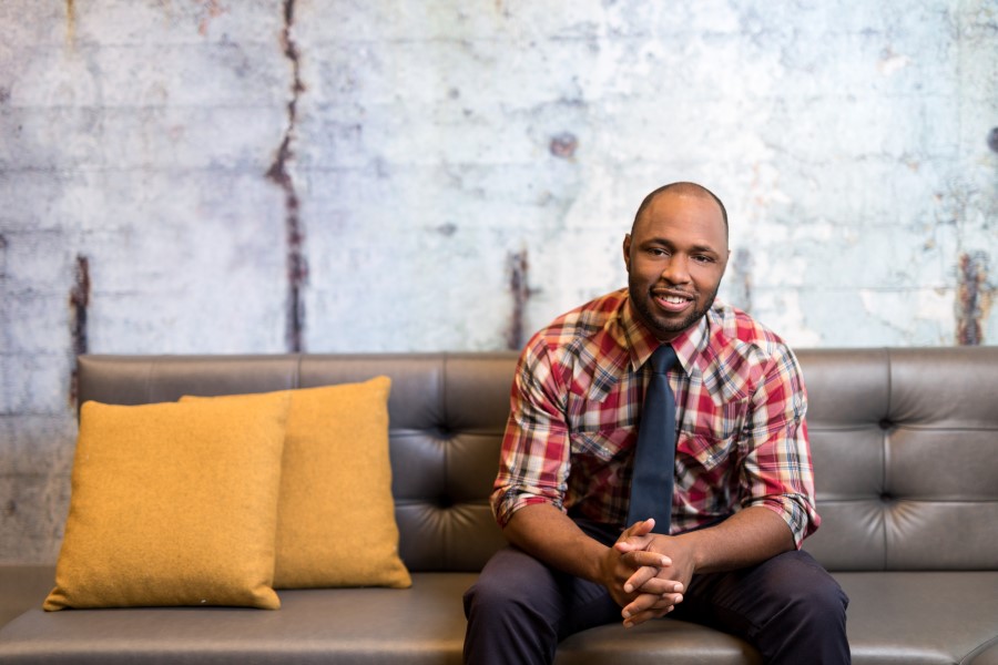 Portrait of man with a tie and plaid button down shirt sitting on a couch facing the camera, smiling with his hands folded.