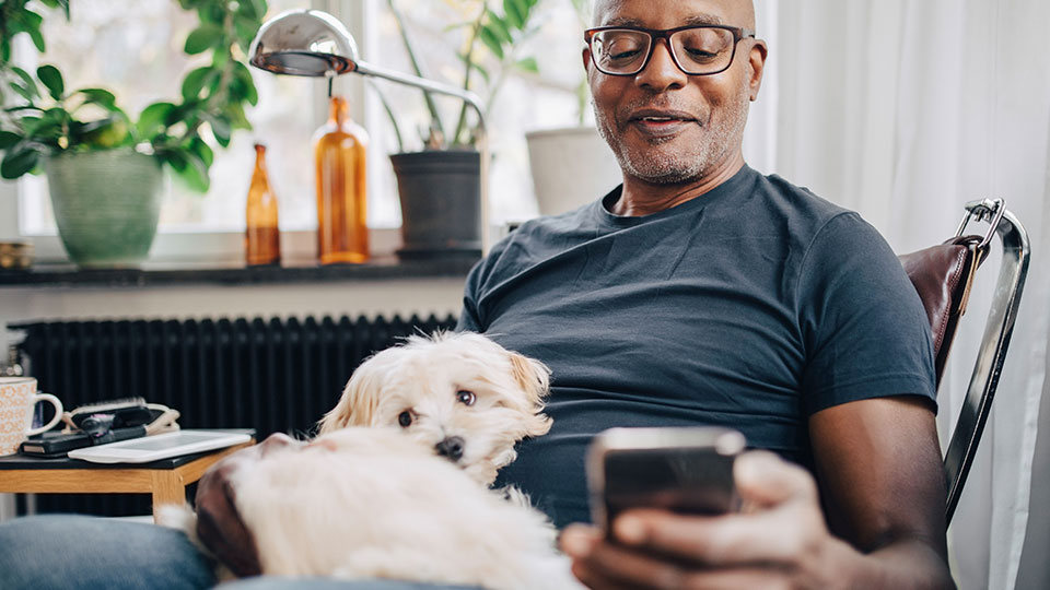 A man in a home environment sits with a dog in his lap while he reads his phone