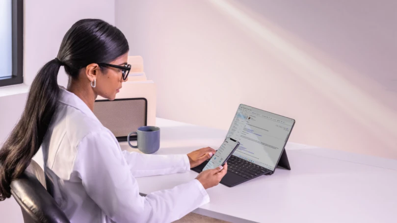 Woman sitting at desk with laptop open and cell phone in hand.