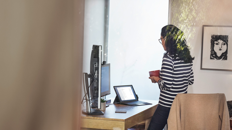 Femme travaillant depuis son bureau &agrave; domicile sur un appareil Surface Pro, debout &agrave; son bureau avec une tasse dans les mains