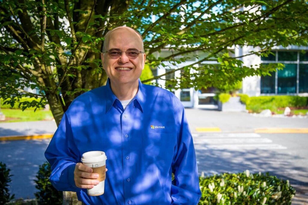 Apple smiles as he stands outside a Microsoft building holding a cup of coffee. 
