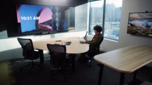 A Microsoft employee is shown working in a conference room with chairs and table configured around the screen.