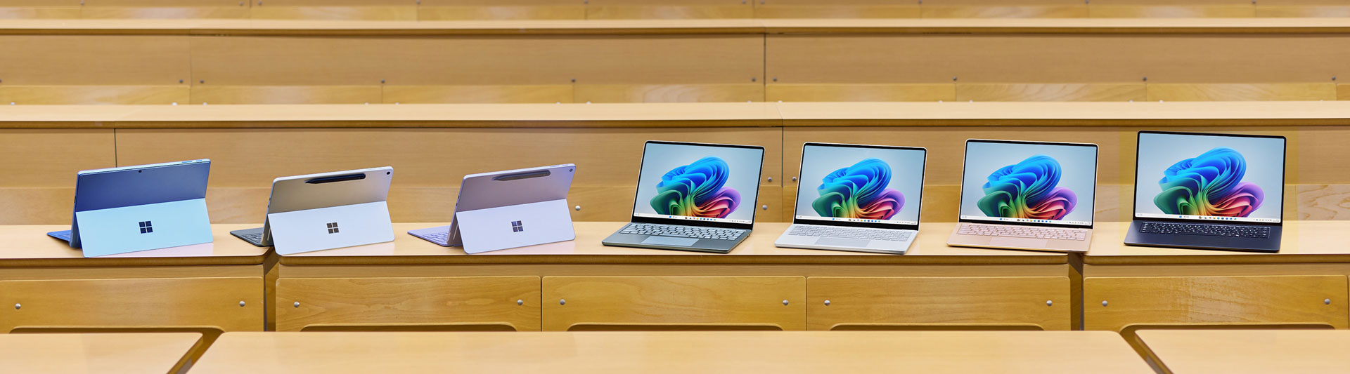 7 台のカラフルな Microsoft Surface タブレットとラップトップが木製の机の上に並べられており、開いたラップトップには Windows 11 の壁紙が表示されています。