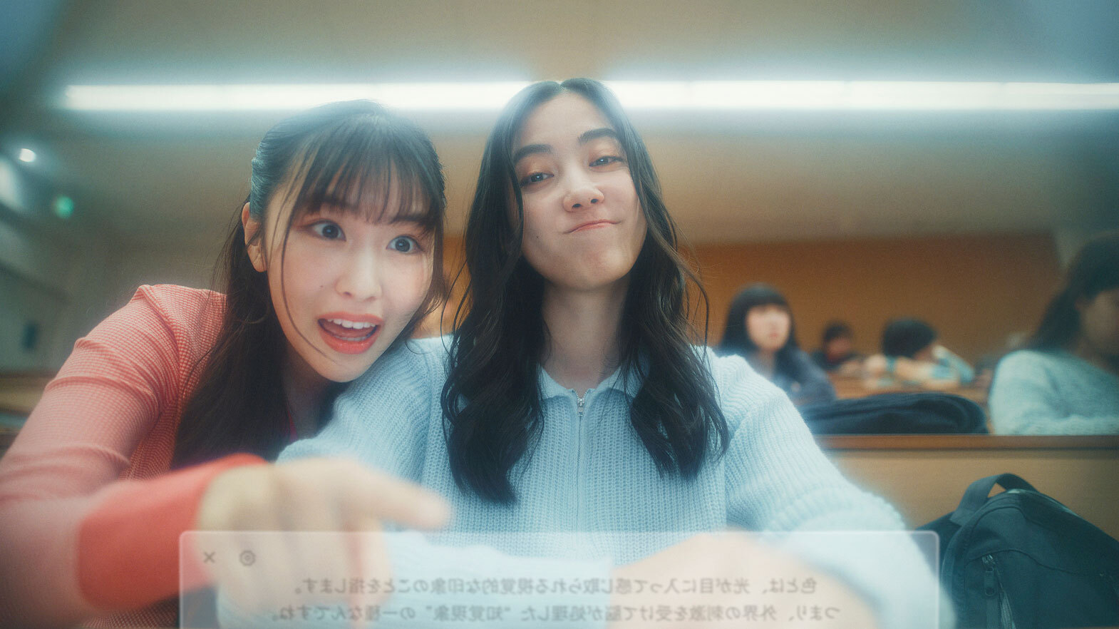 講義室に座っている二人の女子学生。左側の学生は画面上のテキストボックスを指差して驚いた表情を見せ、右側の学生はカメラに向かって遊び心のある笑顔を浮かべている。