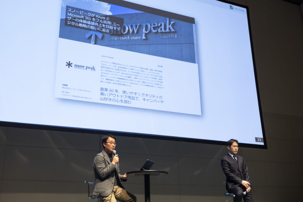 壇上にて大型スクリーンを前に講演を行う男性 2 名