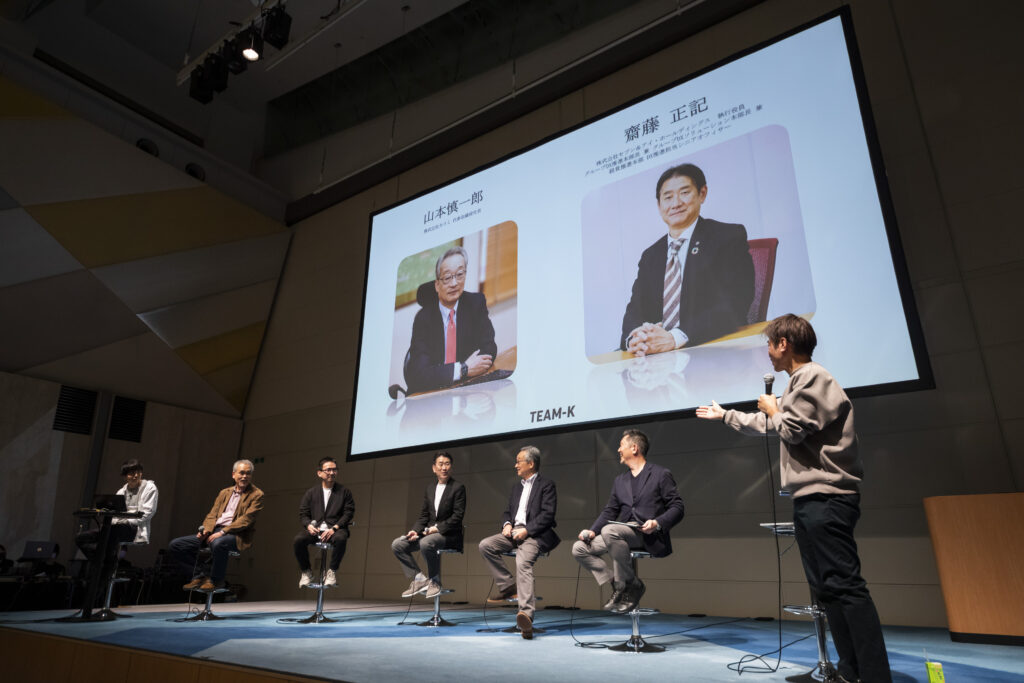 壇上にて大型スクリーンを前に講演を行うパネリスト 6 名と司会者