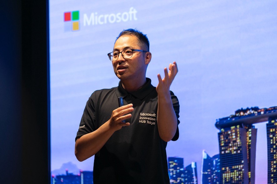 A man in glasses speaking to a man in front of a large screen