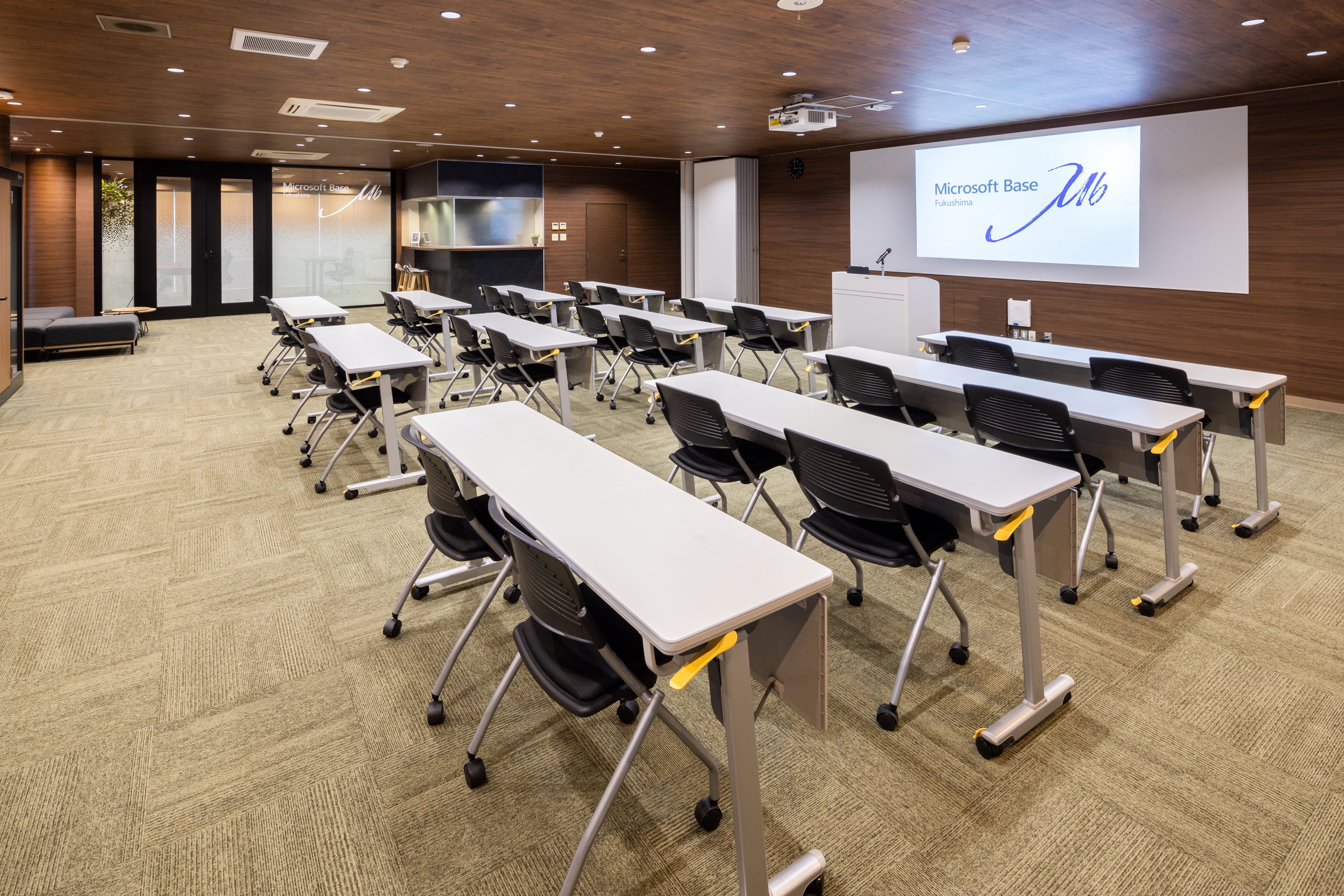 折りたたみ式のデスクと椅子が並ぶセミナー形式の会議室で、プロジェクタースクリーンに「Microsoft Base Fukushima」と表示され、温かみのある木製の壁と天井が特徴です。