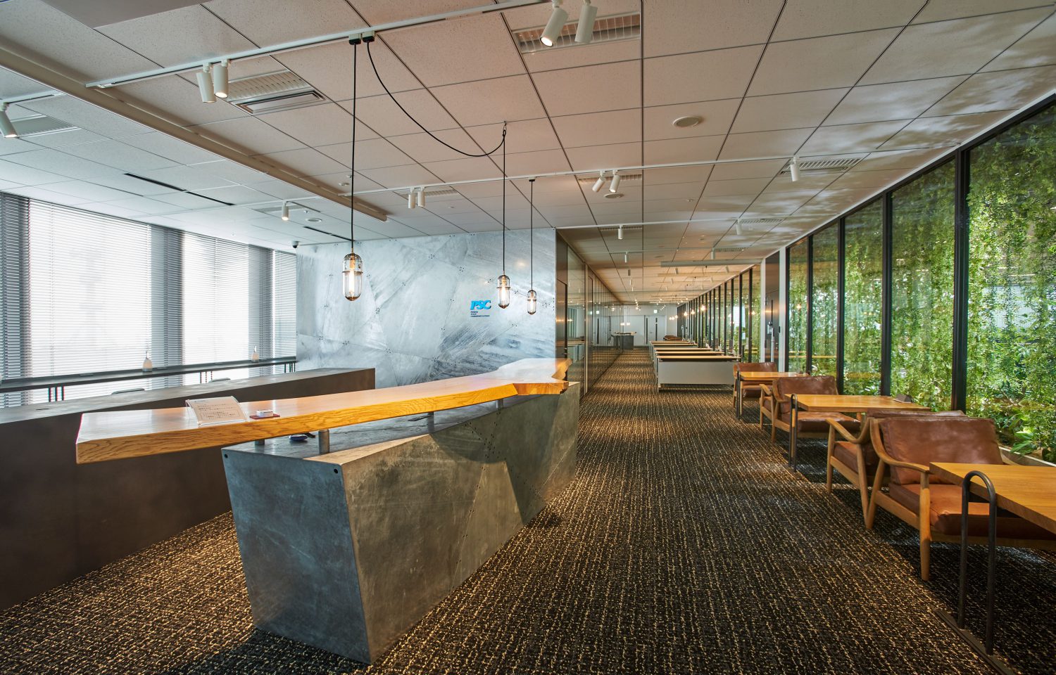 オープンラウンジ。ライブエッジの長い木製カウンターとペンダントライト、植栽のあるガラス面に沿って並ぶレザーアームチェアとテーブル。