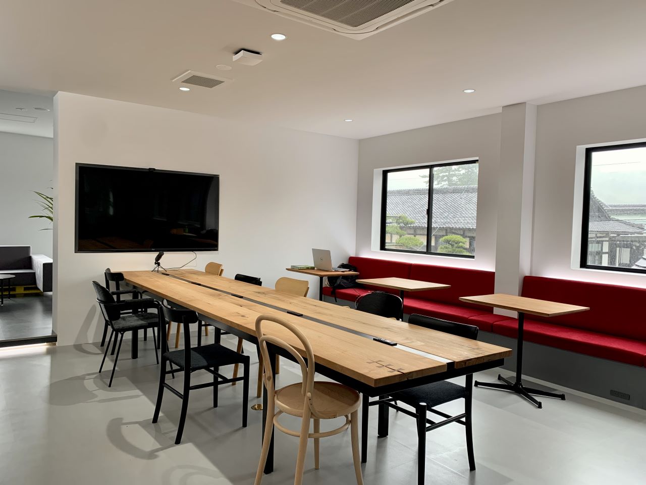 大きな木製の共有テーブルと様々な椅子、窓際の赤いベンチ席、壁掛けディスプレイがある明るい会議スペース。