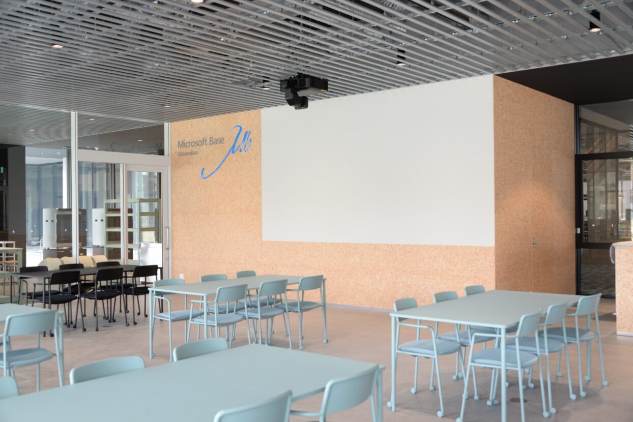 淡いブルーの机と椅子が並ぶ明るい多目的室、大型投影用の壁と近くの「Microsoft Base」ロゴ。