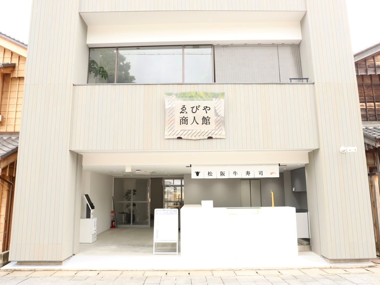 「ゑびや商人館」と書かれた布看板と、入口に「松阪牛寿司」の店舗看板がある白いモダンな2階建ての建物で、周囲には日本の伝統的な木造建築が並んでいます。